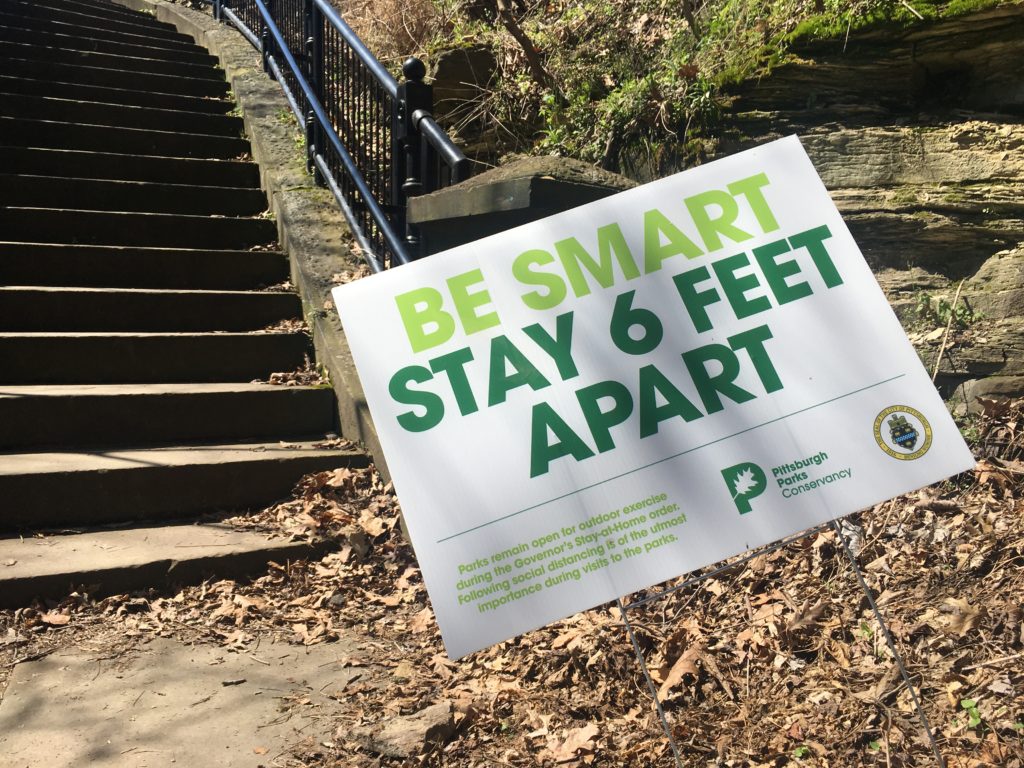 Yard sign at the base of a stairway in a park: "Be Smart -- Stay 6 Feet Apart"